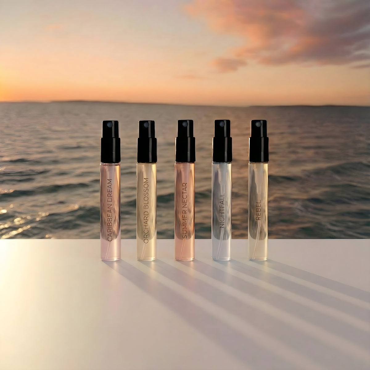 Five spray samples labelled Caribbean Dream, Orchard Blossom, Summer Nectar, Nightfall and Rebel beside the sea at sunset.
